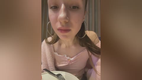 Media: A video of a young woman with light skin, brown hair in pigtails, wearing a pink top and large hoop earrings, sitting at a table with a fork in a white plate.