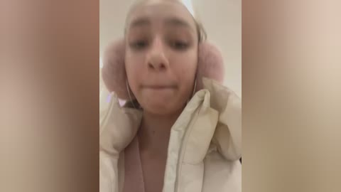 Media: A blurry video of a young woman with light skin and blonde hair, wearing a white and beige puffer jacket, standing in a dimly lit room. Her ears are covered with large, fluffy pink earmuffs.