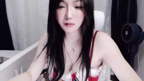 Media: Video of a fair-skinned woman with long black hair, wearing a red and white striped bra, sitting in front of a white bathtub, with a fan and a white chair in the background.