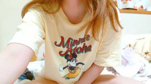 Media: Video of a woman with light skin and long red hair wearing a yellow t-shirt with a Mickey Mouse design, sitting on a bed with white sheets and pillows.