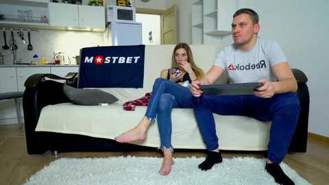 Media: Video of a man and woman playing a video game on a couch in a modern, minimalist living room with white walls, a white kitchen in the background, and a fluffy white rug.