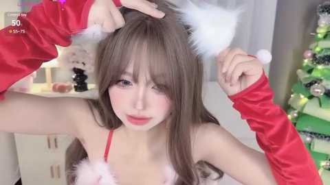 Media: Video of a young Asian woman with long brown hair, wearing a red and white Santa hat, adjusting her fur ears, in a festive room with a decorated Christmas tree.