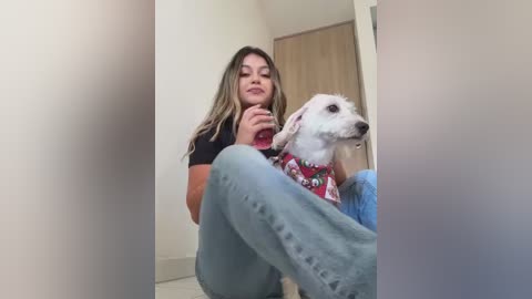 Media: Video of a young woman with long, wavy brown hair, wearing a black t-shirt and blue jeans, sitting on the floor, holding a small white dog.