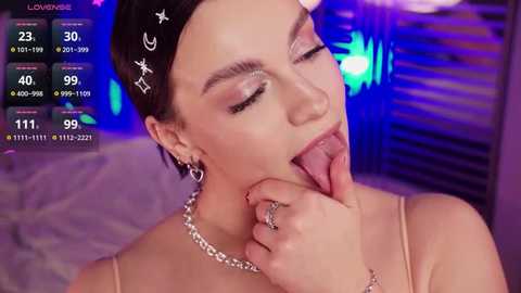 Media: Video of a woman with short dark hair, wearing a silver chain necklace and a star headband, licking her lips, set against a blurred, purple-lit background.