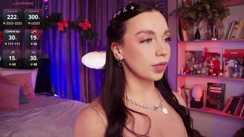 Media: Video of a young woman with long, dark hair, wearing a black headband and silver necklace, standing in a cozy, dimly-lit room with holiday decorations, shelves filled with books, and a bed in the background.