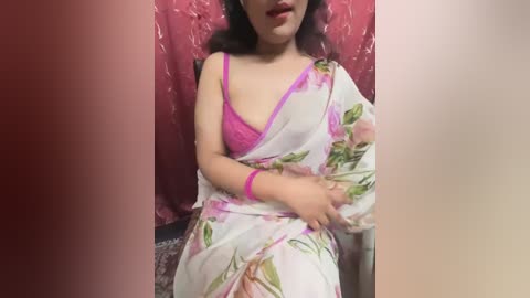 Media: A video of an Indian woman with medium skin tone and dark hair, wearing a pink bra and white floral saree, seated against a red, textured wall.