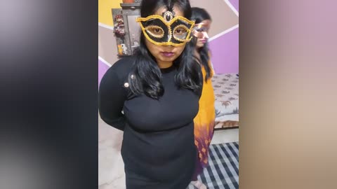 Media: Video of a woman with medium skin tone and black hair, wearing a black dress and a yellow and black masquerade mask, standing in a room with purple and beige walls.