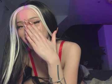 Media: Video of a young woman with long, straight, black hair with white streaks, wearing a red bra. She covers her face with one hand, revealing a tattoo on her wrist. Dim, purple-lit room in the background.