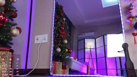 Media: Video of a modern bedroom with a Christmas tree adorned with red and gold ornaments, a white bed, and a room divider with a glowing lamp, under purple LED lights.