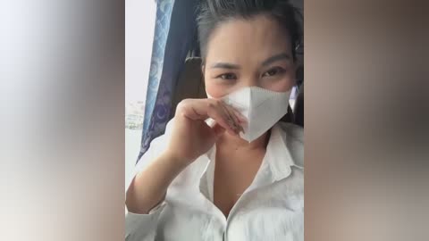 Media: Video of an Asian woman with medium skin tone, wearing a white shirt, holding a tissue to her face, in a bus with blue curtains.