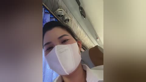 Media: A close-up video of a woman with long dark hair wearing a white surgical mask, looking directly into the camera. The background features a ceiling with exposed ductwork and fluorescent lights.