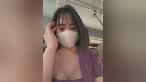 A video of an East Asian woman with shoulder-length dark hair, wearing a white surgical mask, a purple top, and a distressed expression, taken in a dimly lit room with a ceiling fan.