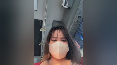 Media: Video of a woman with medium-length brown hair and a surgical mask, wearing a red top, standing in a hospital room with medical equipment and a curtain in the background.