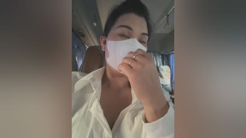 Media: Video of a woman wearing a white surgical mask, white shirt, and sitting in a bus with dim lighting. She appears to be coughing into her hand, which is holding a tissue.