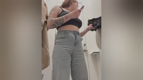 Media: Video of a slim, tattooed woman in a black sports bra and high-waisted, light-wash jeans, holding a black shirt in a changing room.