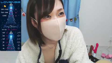 Media: Video of an Asian woman with shoulder-length black hair, wearing a face mask, a beige sweater, and a necklace, in a room decorated with blue curtains and a \"LOVENSE\" sign.