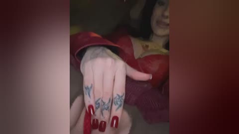 Media: Video of a woman with pale skin, long dark hair, and red lipstick, wearing a red leather jacket, holding her hands with blue and green tattoos. Background is blurry, indoor setting.