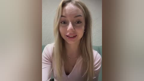 Media: Video of a smiling young woman with long blonde hair, fair skin, and a pink top, seated against a beige wall.