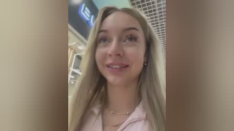 Media: Video of a young woman with long blonde hair, wearing a pink shirt, smiling. She's in a bathroom with tiled walls and a mirror.