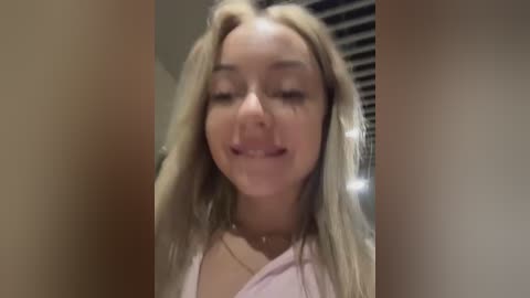 Media: Video of a smiling young woman with long blonde hair, wearing a pink shirt, standing in a dimly lit room with vertical blinds.