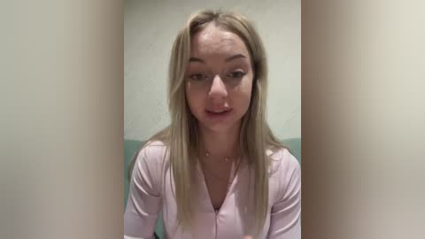 Media: A video of a young, fair-skinned woman with long, straight blonde hair, wearing glasses and a pink top, standing in front of a light green wall.