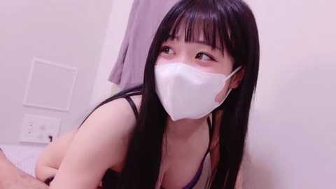 Media: A video of an Asian woman with long black hair and a white mask, wearing a purple bra, kneeling on a bed with white sheets, in a simple bedroom setting.