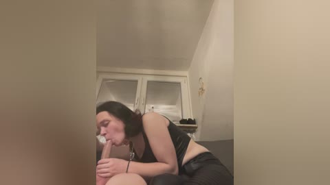 Media: Video of a fair-skinned woman with dark hair performing oral sex on a man in a dimly-lit bedroom with beige walls and a white-framed window.