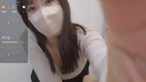 Media: Video of an East Asian woman with long black hair, wearing a white face mask, a white ribbed sweater, and a black skirt, captured indoors with blurred background.