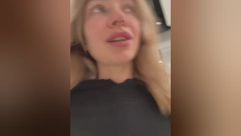 Media: A blurred video of a woman with light skin, blonde hair, and a septum piercing, wearing a black top, seen from the waist up. The background is out of focus.