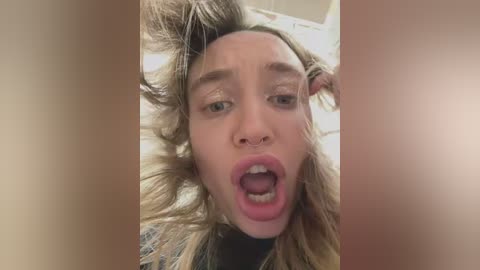 Media: Video of a young woman with curly blonde hair, distressed facial expression, mouth open wide, wearing a black top, indoors with blurred background.