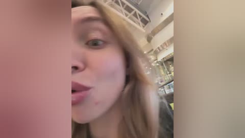 Media: A blurry, close-up video of a Caucasian woman with blonde hair, wearing pink lipstick, and a dark shirt, standing in a dimly lit, modern store with shelves in the background.