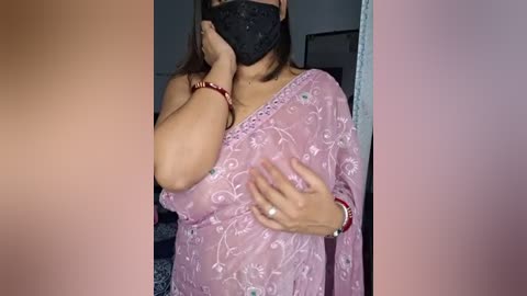 Media: Video of a South Asian woman in a pink saree, wearing a black mask, and covering her mouth with her hand. Background includes a blurred wall and furniture.