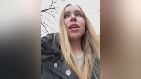 Media: A video of a young Caucasian woman with long blonde hair, wearing a dark jacket, taken outdoors with bare tree branches in the background.