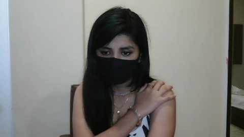 Media: Video of a young woman with long black hair, wearing a black mask, white top, and multiple necklaces, seated against a plain white wall, with a glimpse of a room in the background.