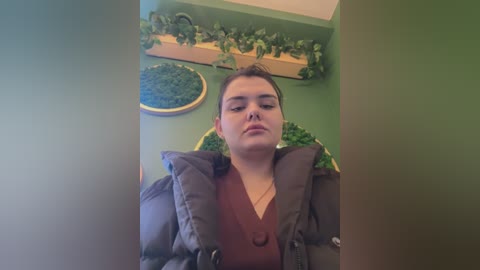 Media: Video of a young, fair-skinned woman with short brown hair, wearing a brown puffer jacket, sitting in a green room with a wooden shelf and greenery.