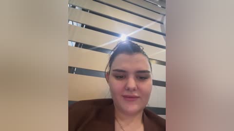 Media: Video of a young woman with light skin, brown hair tied back, wearing a brown blazer, standing in front of modern, beige and metallic vertical blinds.