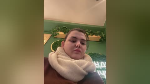 Media: Video of a young woman with light skin, dark hair, and a white scarf, standing indoors with a green wall and potted plants in the background.