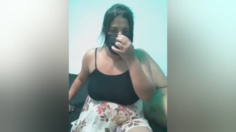 Media: A video of a plus-sized woman with medium skin tone, wearing a black tank top and floral skirt, sitting in a dimly lit room with a blurred background.