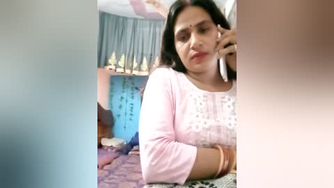 Media: Video of a woman with medium skin tone, dark hair, wearing a pink embroidered kurta, talking on a phone, in a colorful, decorated room with blue walls and Hindu religious symbols.