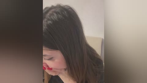 Media: A video of a young woman with long, straight, dark brown hair, wearing red lipstick, sitting in a beige chair against a plain, light-colored wall.