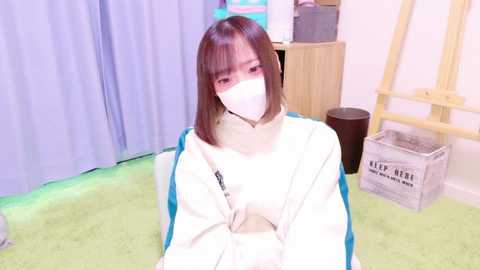 Media: Video of an Asian woman with straight brown hair and a white mask, wearing a white and blue hoodie, sitting on a green carpet. Background features a light-colored room with a wooden stool and a \"KID NERD\" box.