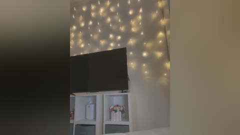 Media: Video of a cozy room with a large, flat-screen TV mounted on a beige wall adorned with warm, golden fairy lights. Below, white shelves hold decorative items, including a bouquet of flowers and a framed picture.