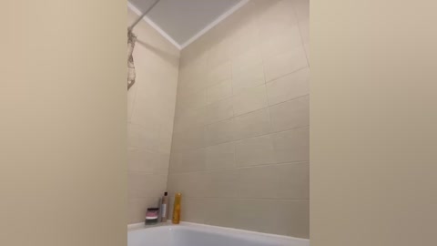 Media: A video of a simple, clean bathroom featuring a beige tiled shower stall with a white bathtub and a single bottle of soap on the ledge. The walls are uniformly tiled with light beige tiles, and the ceiling has a white border.