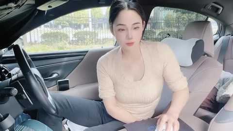 Media: Video of a young East Asian woman with fair skin, dark hair tied back, wearing a beige knit top and dark jeans, sitting in a car with beige leather seats, looking slightly distracted.