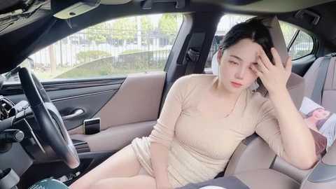 Media: Video of an Asian woman in a beige dress, sitting in a car, looking tired, with a child's toy beside her.