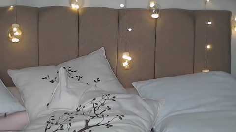 Media: Video of a neatly made bed with beige tufted headboard, white pillows, and a white duvet featuring a black tree pattern. Warm, glowing string lights hang above, adding a cozy ambiance.