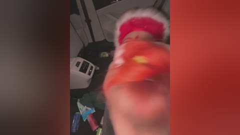 Media: Video of a person wearing a red Santa hat and a bright red jacket, holding a red object, in a messy room with a white microwave and scattered items.