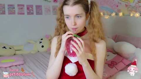 Media: Video of a slender, light-skinned woman with long, curly blonde hair in pigtails, wearing a red Santa dress, drinking from a red wine glass. Background features plush toys, festive decor, and a bed with pink sheets.