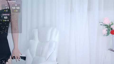 Media: Video of a woman in a black dress with white polka dots, holding a remote control, standing beside a white armchair in a minimalist room with white curtains and a vase of pink roses.