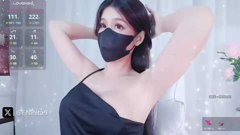 Media: Video of an East Asian woman with long black hair, wearing a black mask, tank top, and revealing cleavage, arms raised. Background shows a room with a white curtain and a digital display with \"Lovenad\" and \"Xinwita\" text.